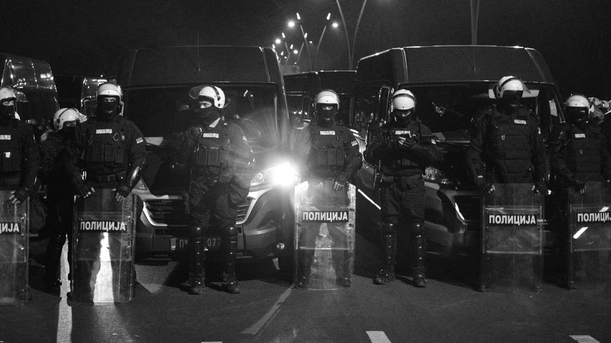 Riot police in protective gear and shields stand in line at night.
